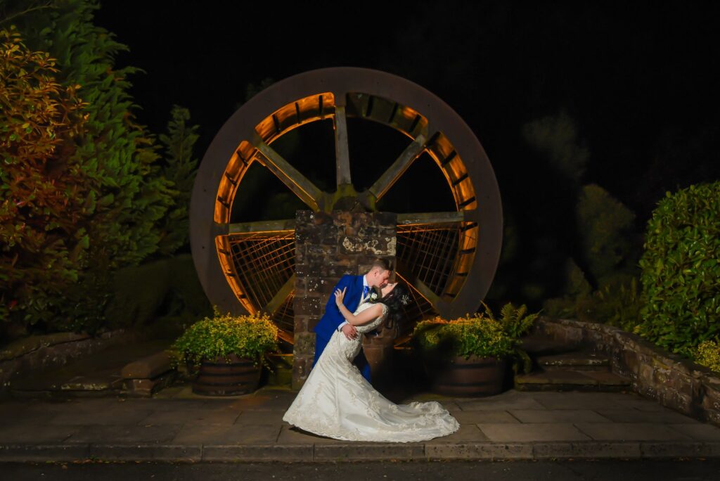 A bride and groom kiss in front of a water wheel at night.
