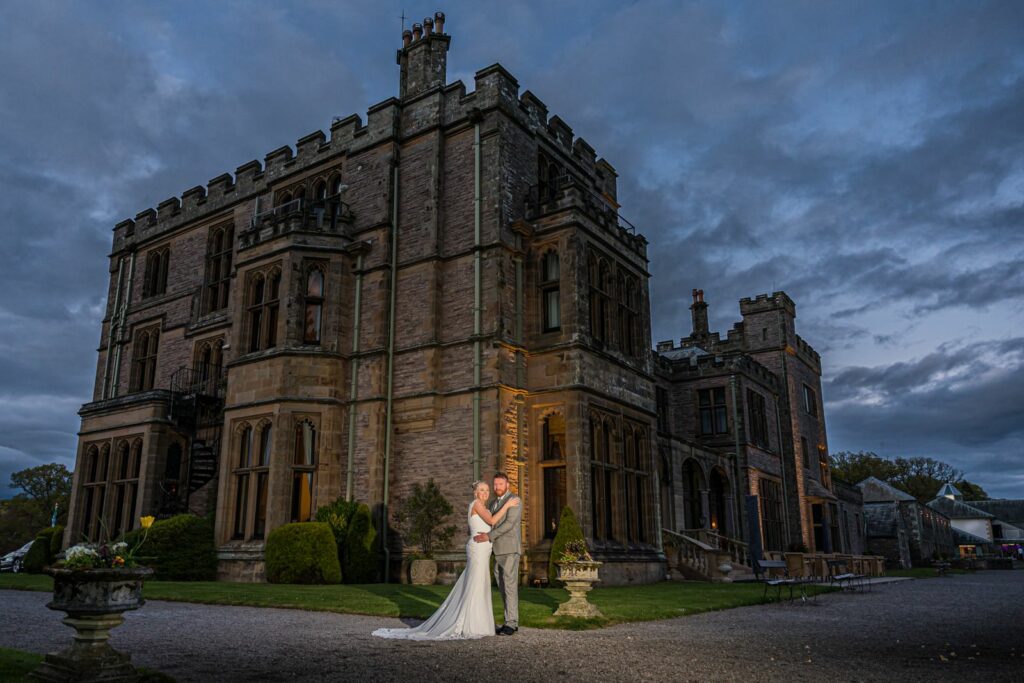 A bride and groom standing in front of a castle at dusk.