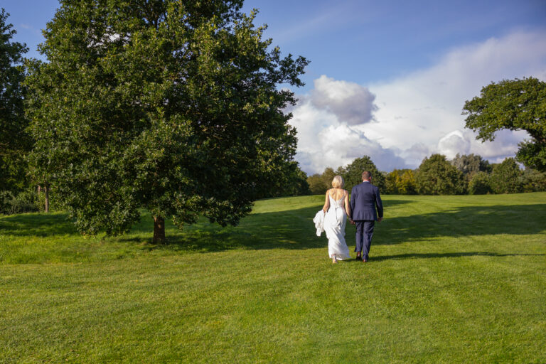 A bride and groom walking through a grassy field.