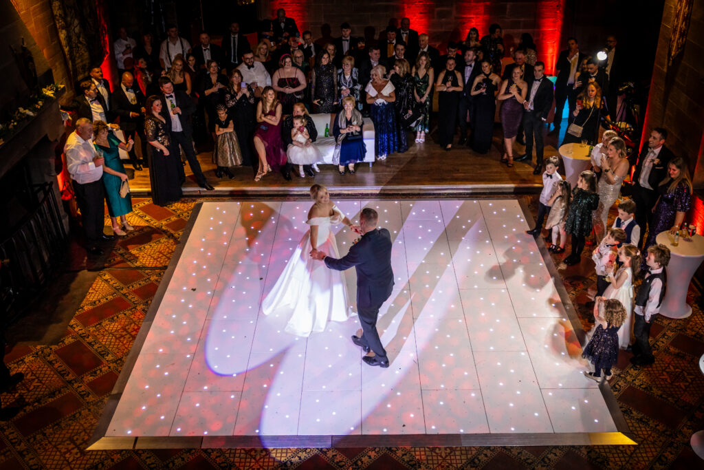 A bride and groom dance on a lit white floor while guests stand around watching at a formal indoor Peckforton Castle Wedding.