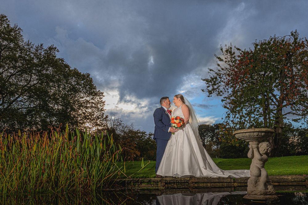 A bride and groom standing in front of a pond under a cloudy sky.