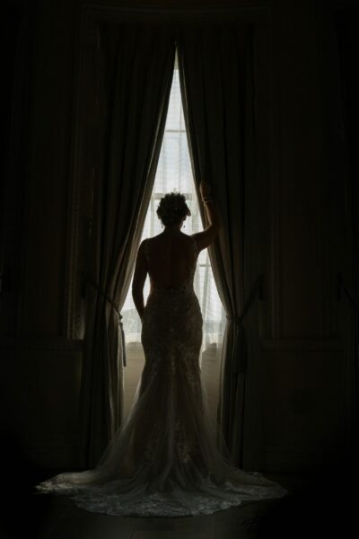 Bride silhouetted against window, elegant dress, romantic setting.