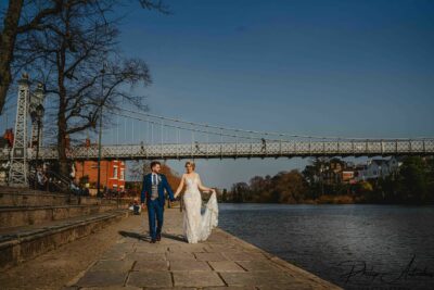 Couple walking by river with bridge in background.