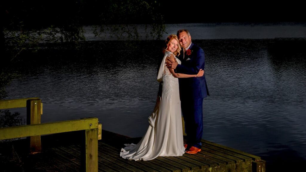 Couple embracing by lake at dusk in wedding attire.