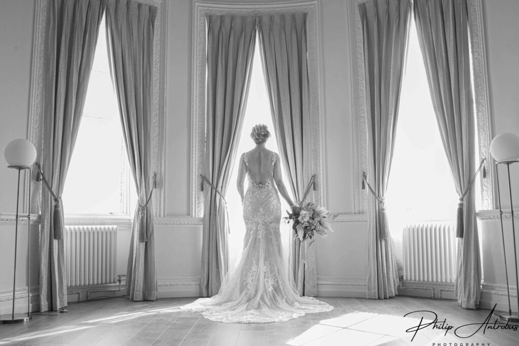 Bride in gown by window, monochrome vintage room.