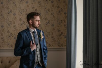 Groom in blue suit by window, thoughtful pose.