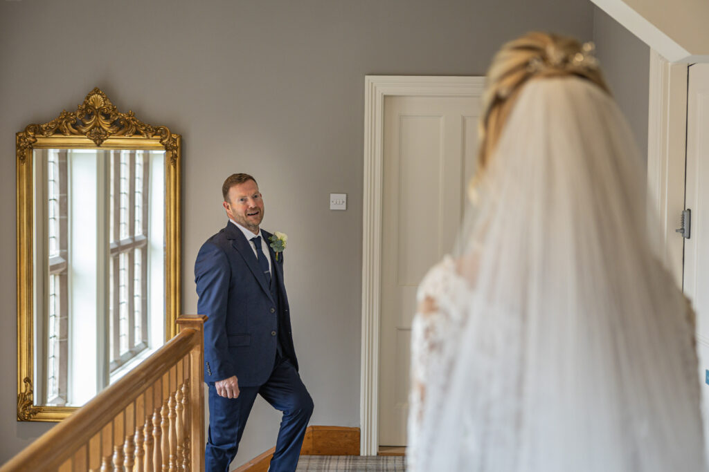 A groom in a navy suit smiles while standing at the top of a staircase, facing a bride in a white dress and veil.