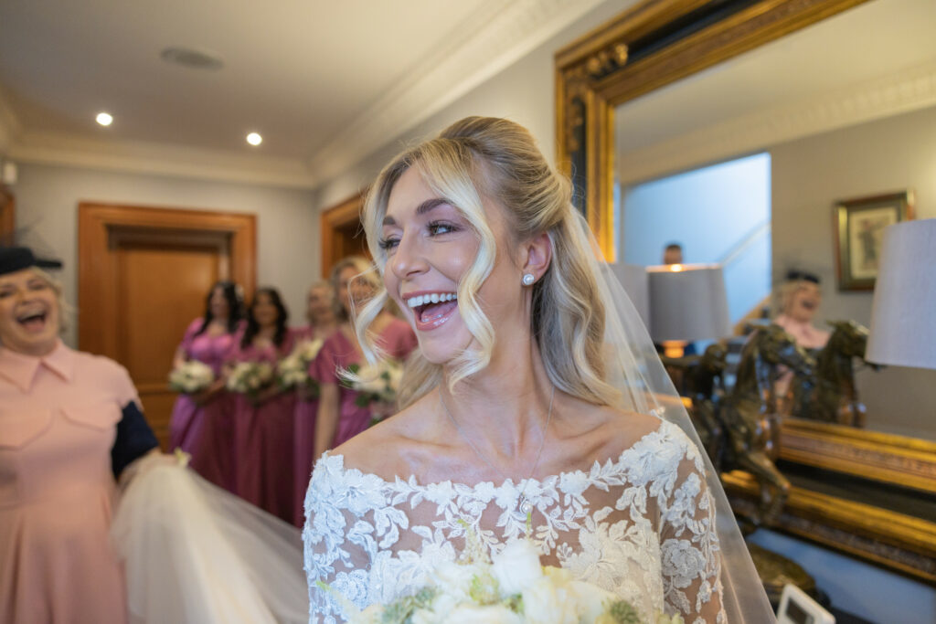 A bride in a lace wedding dress smiles while holding a bouquet, with bridesmaids in pink dresses and a woman in a pink outfit in the background.