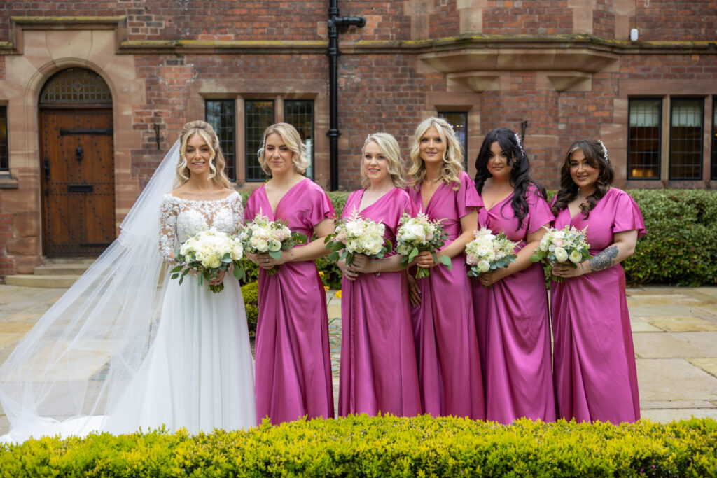 A bride in a white dress stands with five bridesmaids wearing matching magenta dresses, all holding bouquets, in front of a brick building.