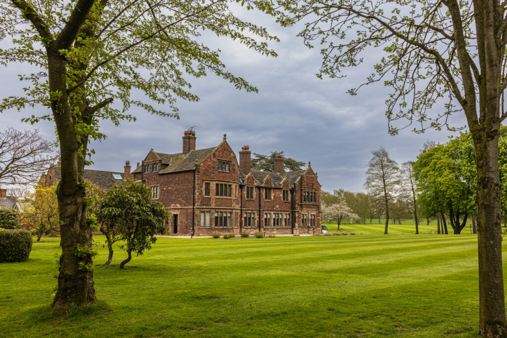 A large brick manor house with multiple chimneys stands on a green, well-maintained lawn, surrounded by trees under a cloudy sky.