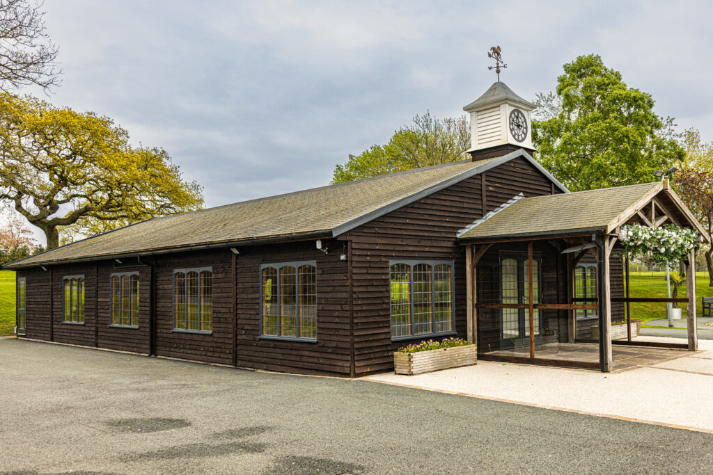 A single-story, dark wooden building with arched windows and a clock tower, surrounded by greenery and a paved area.