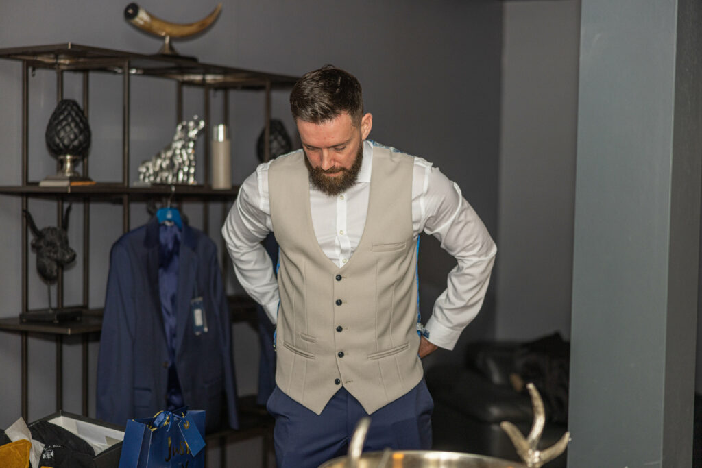 A man with a beard wearing a beige vest over a white shirt stands in a room, looking down with his hands behind his back. A blue suit jacket hangs on a shelf in the background.