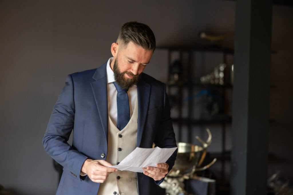 A man in a suit and tie stands indoors, reading a sheet of paper, with shelves and trophies visible in the background.