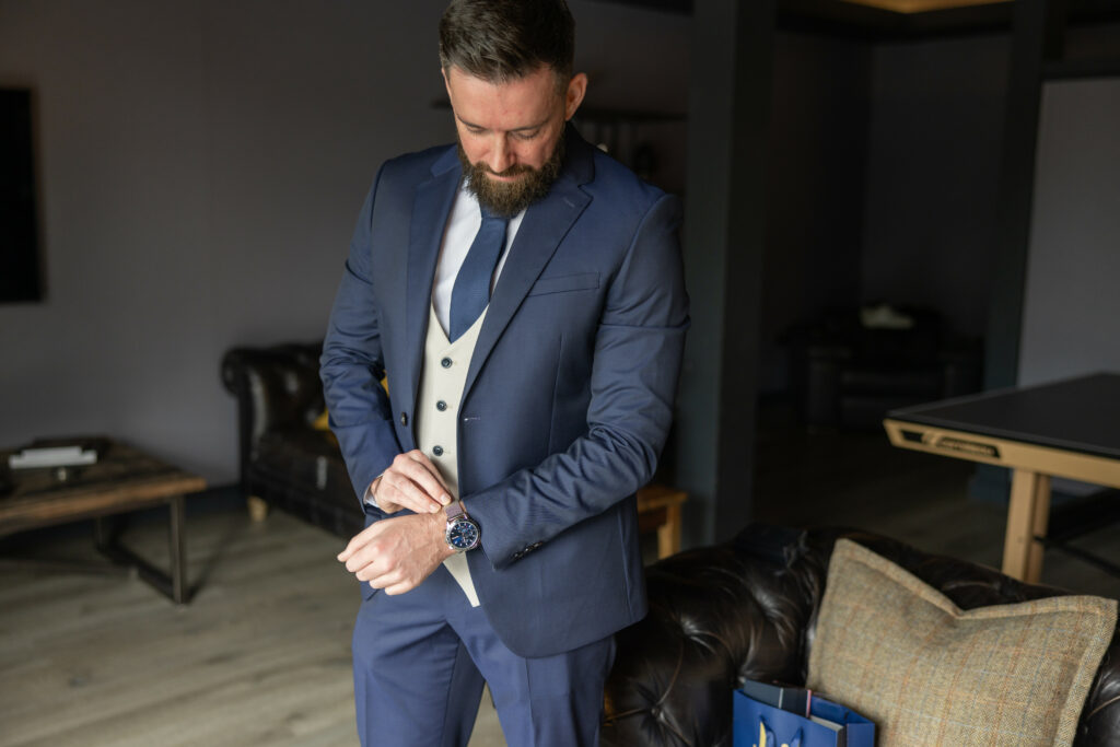 Man in a blue suit and beige vest adjusts his wristwatch while standing in a modern living room with a leather couch and wooden furniture.