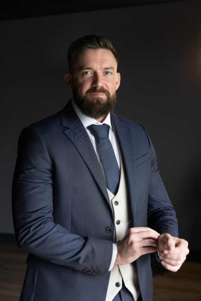 Man with a beard wearing a navy suit, white dress shirt, and blue tie stands indoors adjusting his cufflink, facing the camera with a neutral expression.