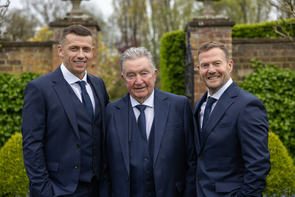 Three men in navy blue suits stand outdoors in front of a garden wall, smiling at the camera.