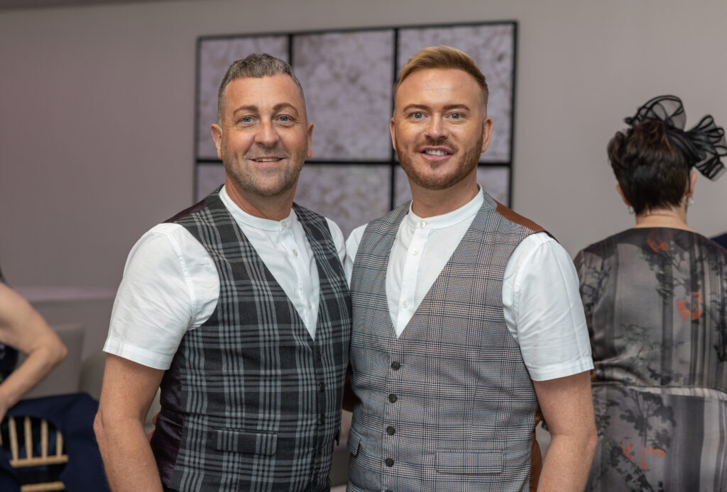 Two men wearing patterned vests and white shirts stand side by side indoors, smiling at the camera. Other people are visible in the background.