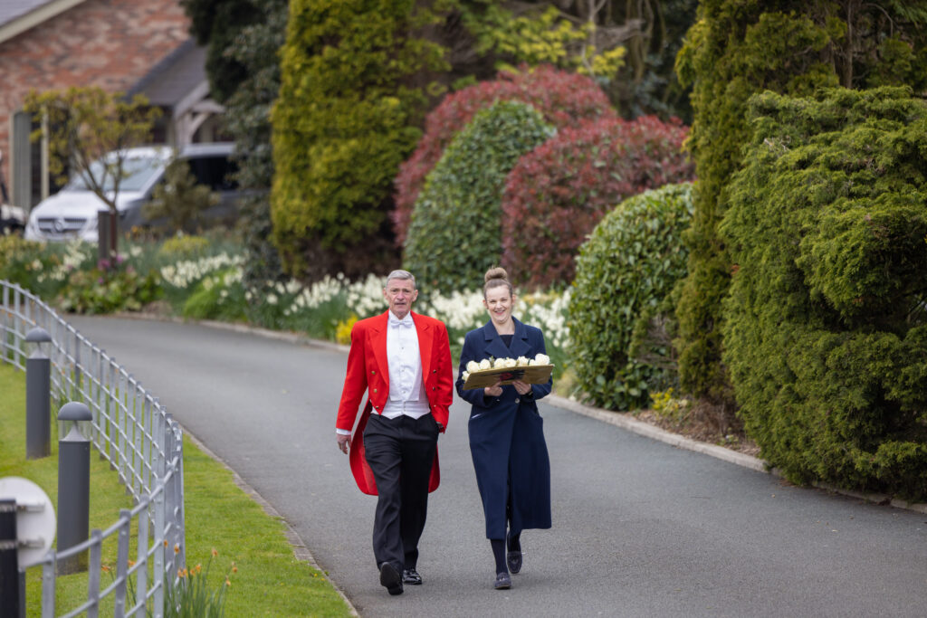 A man in a red tailcoat and a woman carrying a tray of cupcakes walk together along a paved garden path lined with greenery.