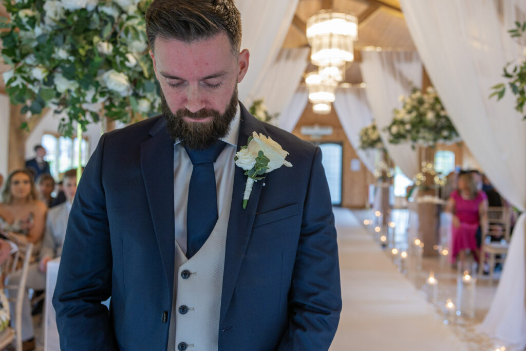 A groom in a navy suit stands with his head bowed, holding a white rose boutonnière, in a decorated indoor wedding venue with white flowers and draped fabric.