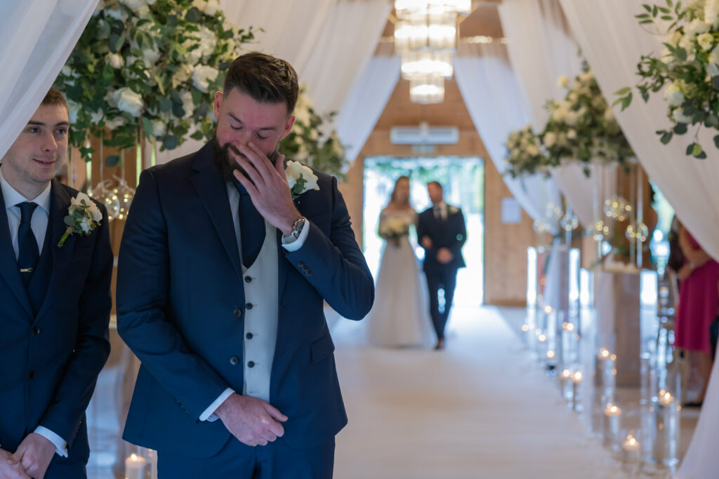 A groom in a navy suit wipes his eyes while standing at the altar as the bride walks down the aisle in the background, accompanied by another person.