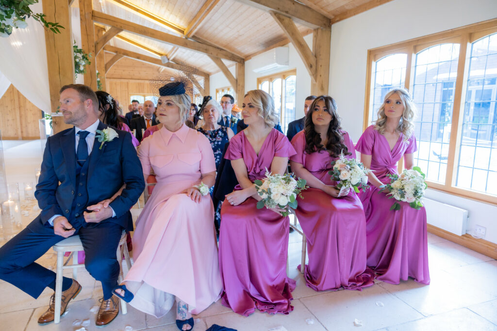 A wedding ceremony with guests seated indoors; three women in matching purple dresses and one in pink sit in the front row holding bouquets.
