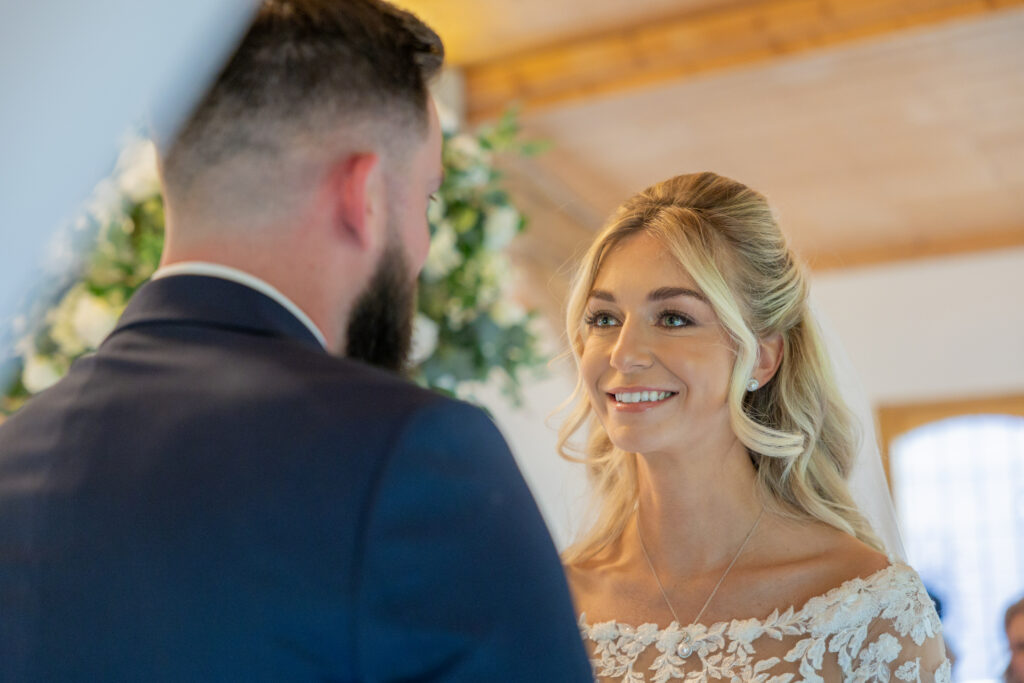 A bride in a white lace dress smiles at a groom in a dark suit during a wedding ceremony indoors.