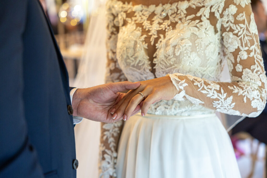Close-up of a groom placing a wedding ring on the bride’s finger during a wedding ceremony. The bride is wearing a white lace dress.