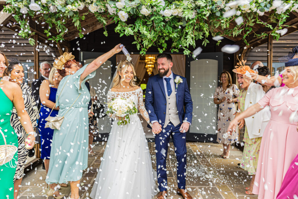 Bride and groom walk beneath flower arch while guests throw confetti, celebrating at an outdoor wedding ceremony.