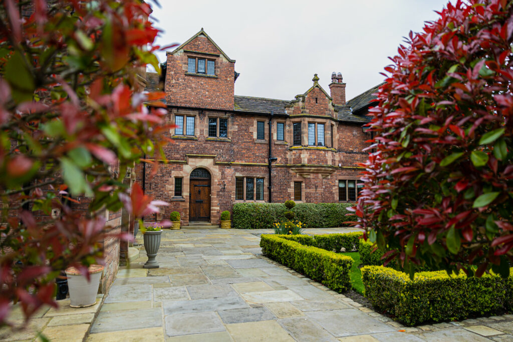 Large brick house with trimmed hedges and potted plants, viewed from a stone walkway framed by red-leaved bushes in the foreground.
