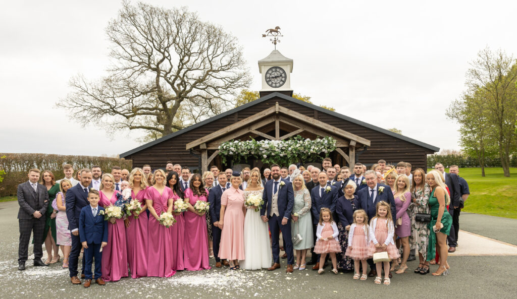 A large wedding group poses outside a wooden chapel, with the bridesmaids in pink dresses and guests of all ages standing together for a formal photo.