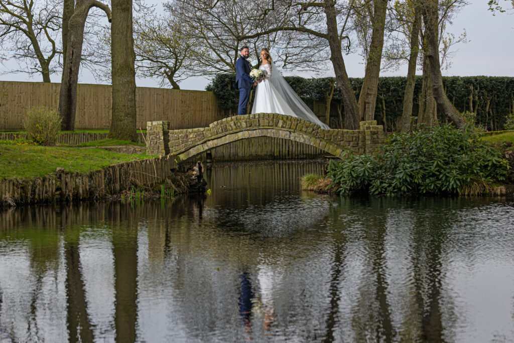 A bride and groom stand together on a stone bridge over a pond, surrounded by trees, with their reflection visible in the water below.