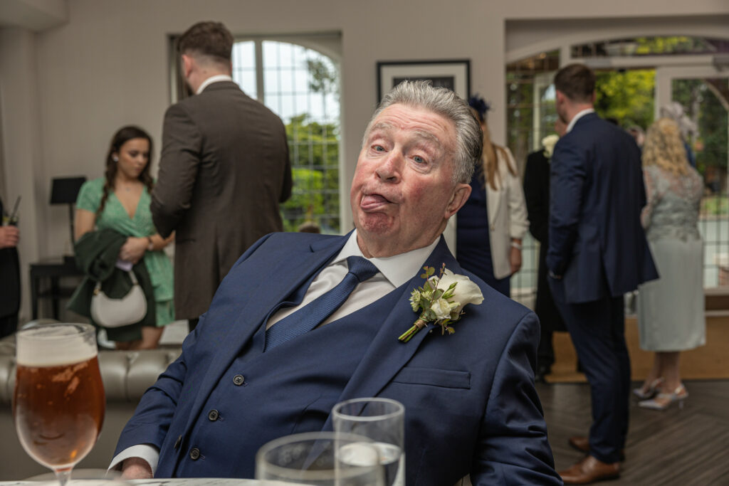 An older man in a suit and boutonniere sits at a table making a funny face; people in formal attire stand and talk in the background.