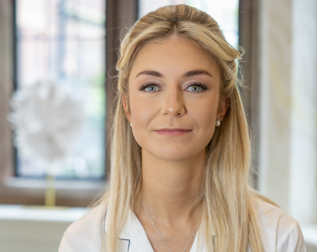 A woman with long blonde hair and light eyes is looking at the camera, standing indoors in front of a window.