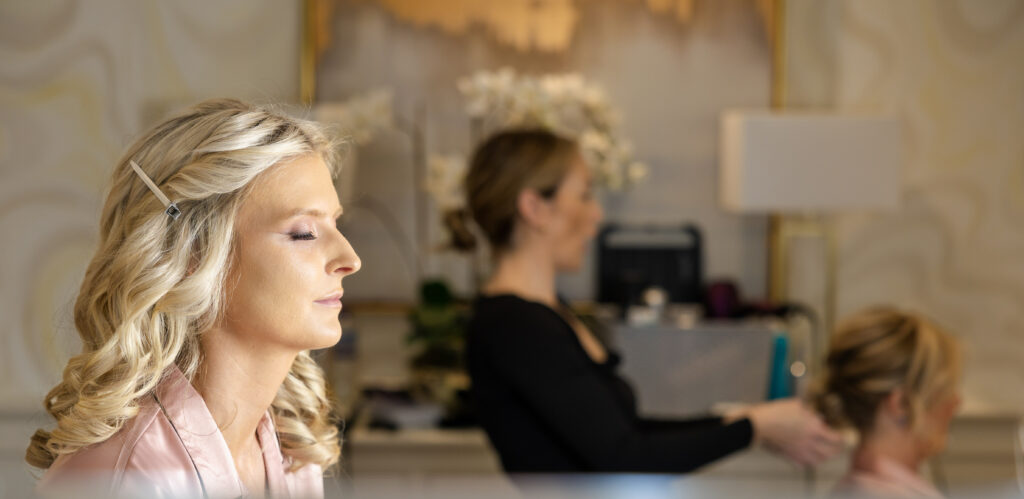 A woman with curled blond hair and hairpins sits with closed eyes, while another woman styles the hair of a third person in the background. The setting appears to be a salon or similar space.
