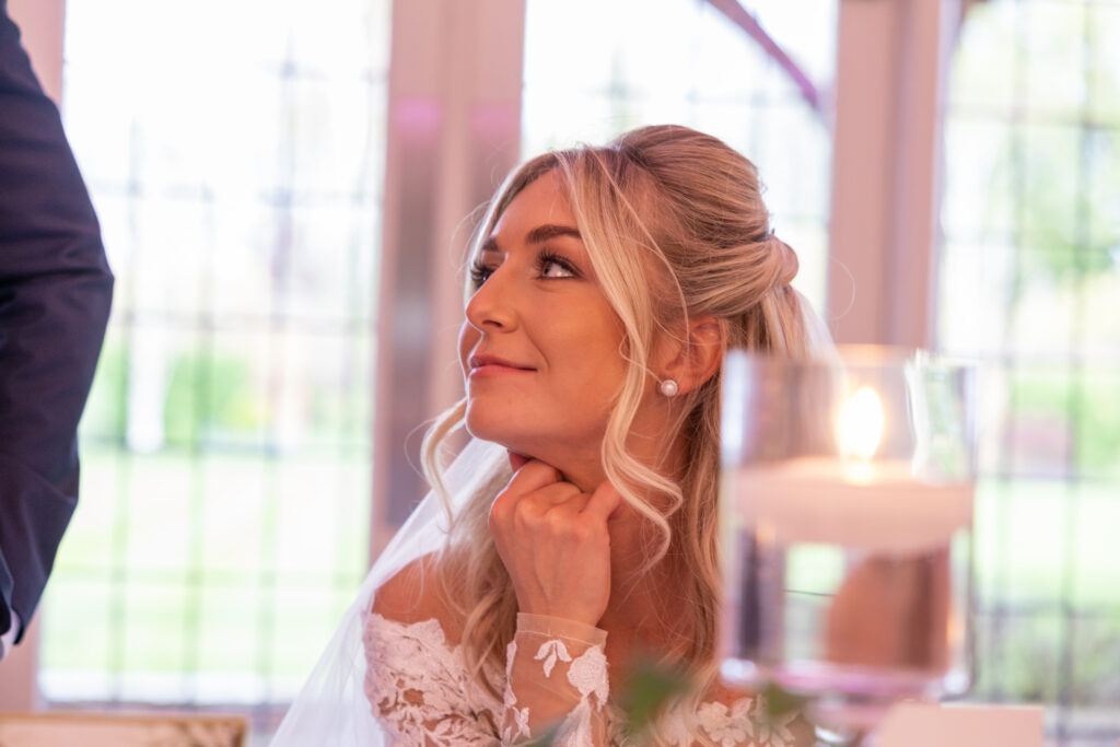 A bride in a white dress and veil sits indoors, resting her chin on her hand and looking upward, with a lit candle in the foreground.