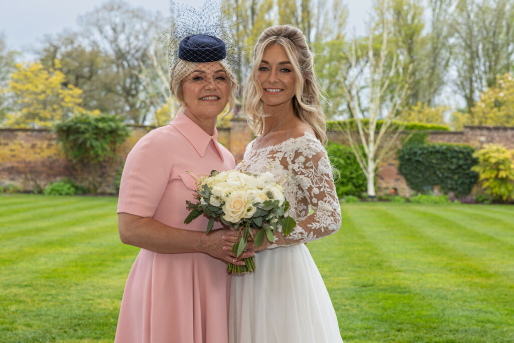 Two women, one in a wedding dress holding a bouquet and the other in a pink dress and hat, stand together outdoors on a green lawn.