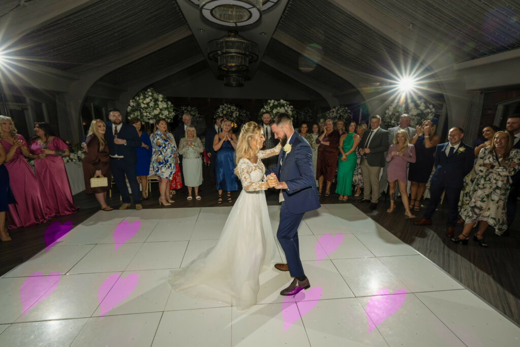 A bride and groom dance together on a white floor with pink heart lights, surrounded by guests in formal attire at a wedding reception.