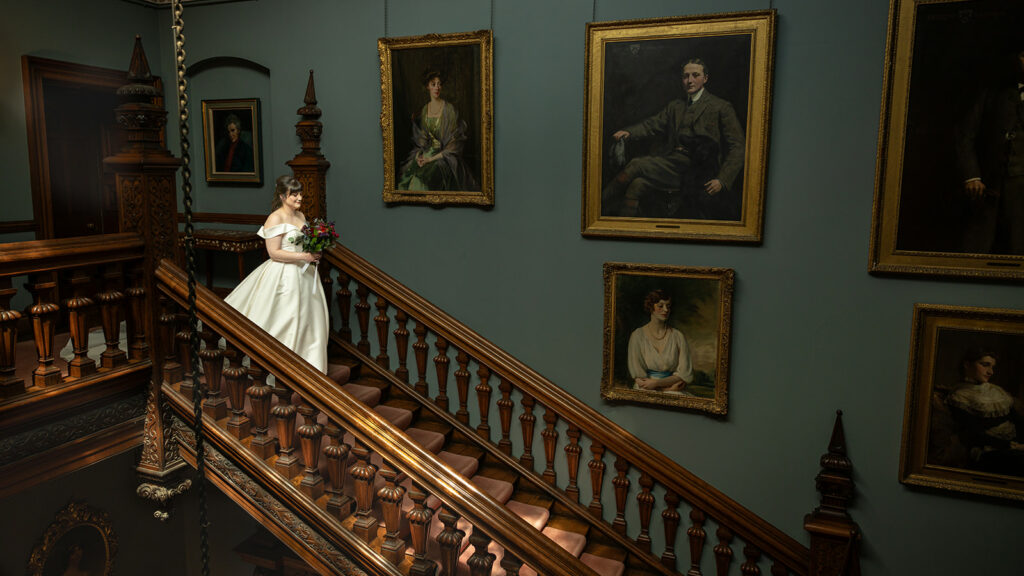 A bride in a white wedding dress ascends a wooden staircase at Arley Hall Wedding, surrounded by intricate wooden detailing and large, framed portraits. She holds a bouquet, embodying elegance and grace.
