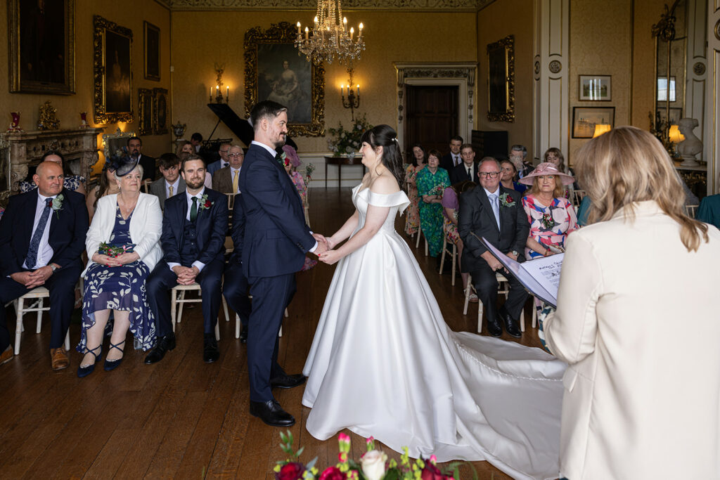 A bride and groom stand holding hands during their Arley Hall Wedding ceremony in a grand room with guests seated on both sides. An officiant stands on the right, reading from a document.