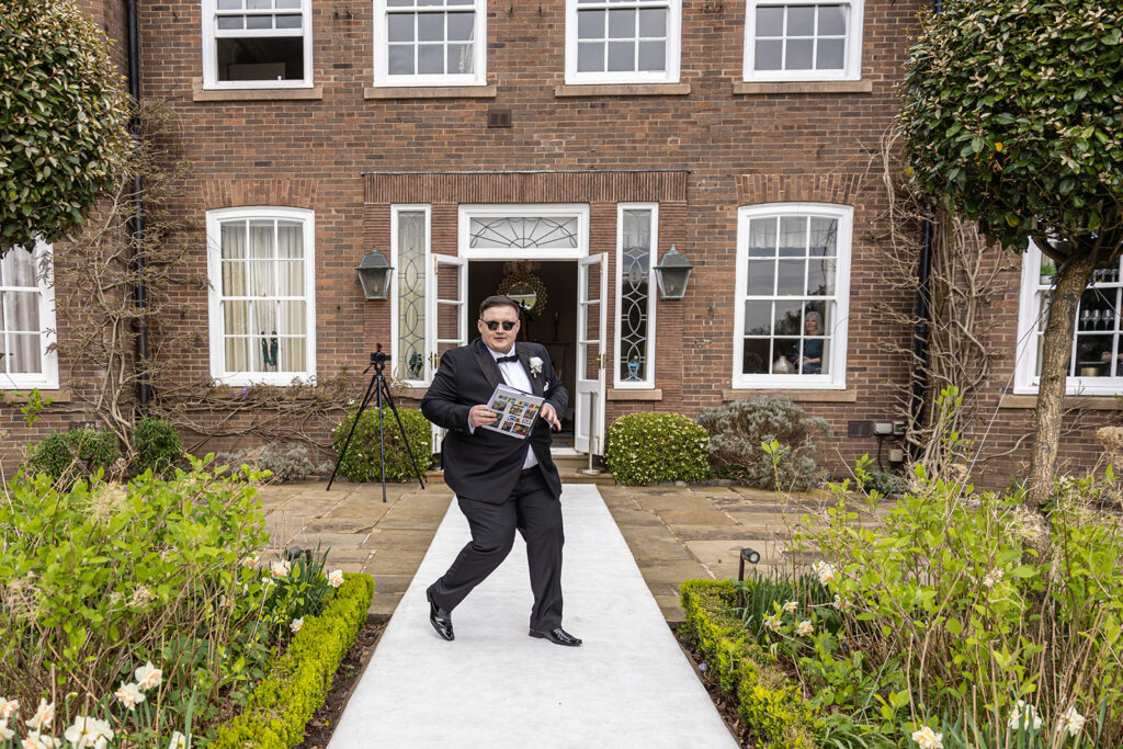 A person in a suit and sunglasses stands on a walkway in front of a brick building at Delamere Manor Wedding, holding a magazine and gesturing with their hand. The building has several windows and is surrounded by greenery.