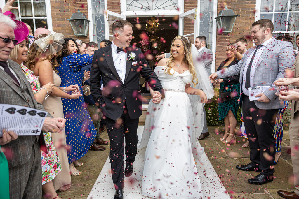 A newlywed couple walking arm in arm down a white aisle while guests throw flower petals in celebration outside Delamere Manor. The groom is in a tuxedo, and the bride is in a white wedding dress, creating a perfect confetti photograph moment.