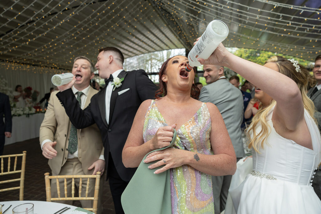 Three people are enthusiastically drinking from bottles in a decorated venue, while others in the background are socializing. The setting appears to be a celebratory event, possibly a wedding. It's moments like these that highlight the joy captured by Cheshire Wedding Photography.