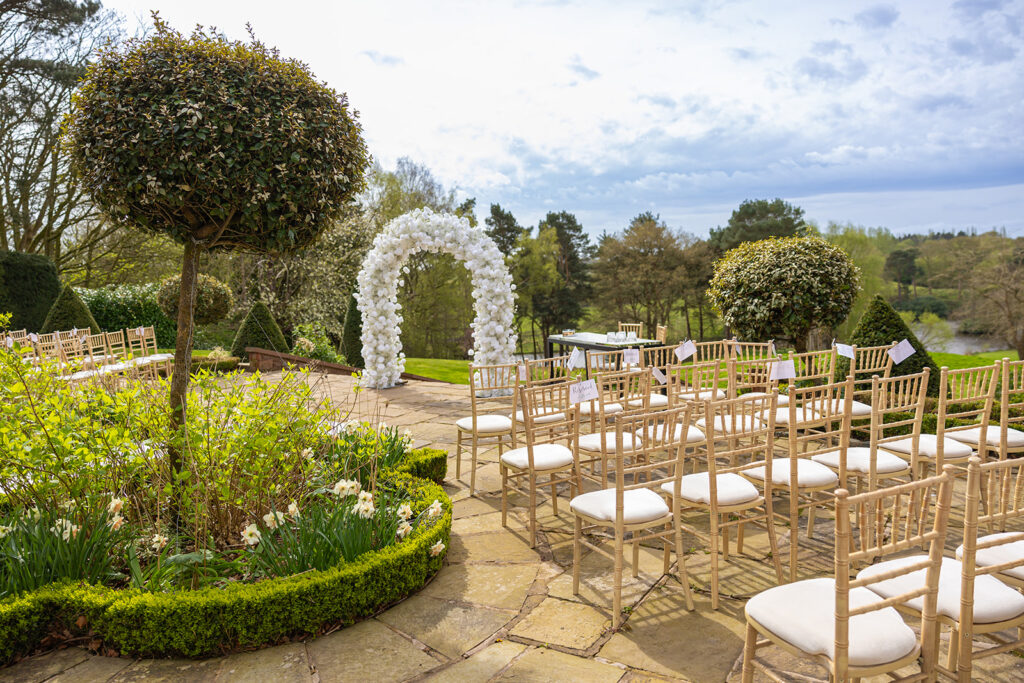 An outdoor wedding setup with rows of white chairs facing a white floral arch. The scene is surrounded by greenery and a stone patio, with a tree and bushes in the foreground, capturing the essence of Delamere Manor Wedding Photography.