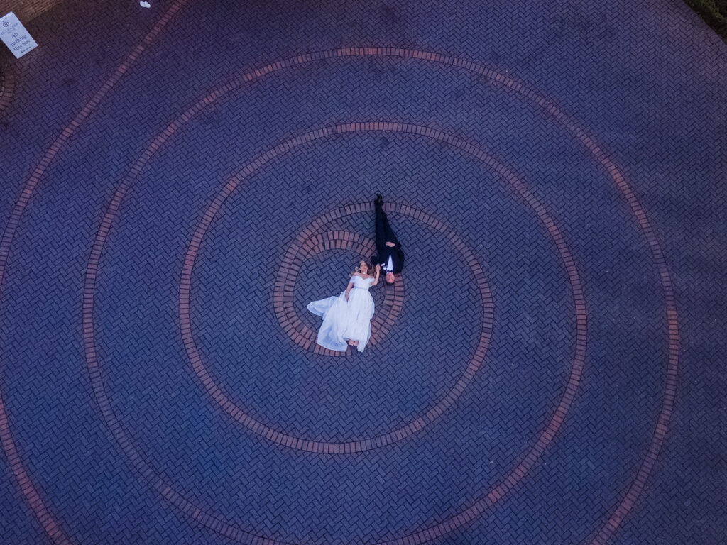 A bride and groom lie on a brick-paved spiral pattern at Delamere Manor, viewed from above through stunning drone photography. The bride dazzles in her white gown while the groom looks sharp in his black suit.