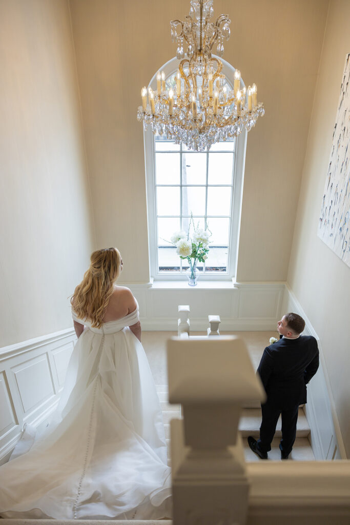 A bride in a white dress stands on a staircase landing, facing away. A groom in a dark suit stands below her, looking up. They are in the well-lit, elegant interior of Delamere Manor, adorned with a large window and chandelier.
