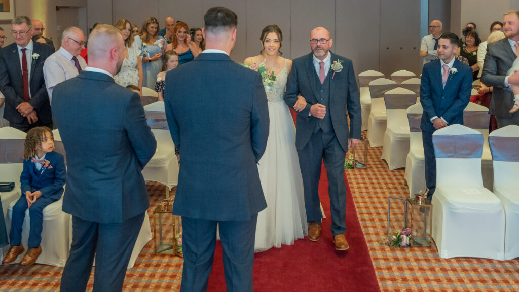 A bride walks down the aisle in Forest Hills with an older man in a suit, approaching two men standing at the altar. Guests are seated on either side.