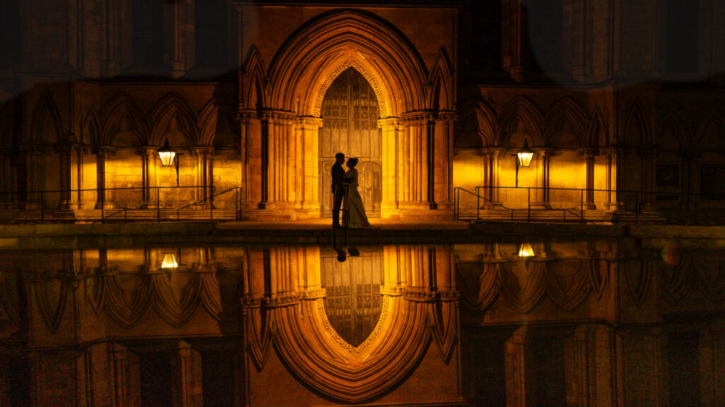 Silhouetted couple stands in front of a lit arched doorway at night, their reflection shimmering in the calm water below, reminiscent of York Cathedral's majestic allure.