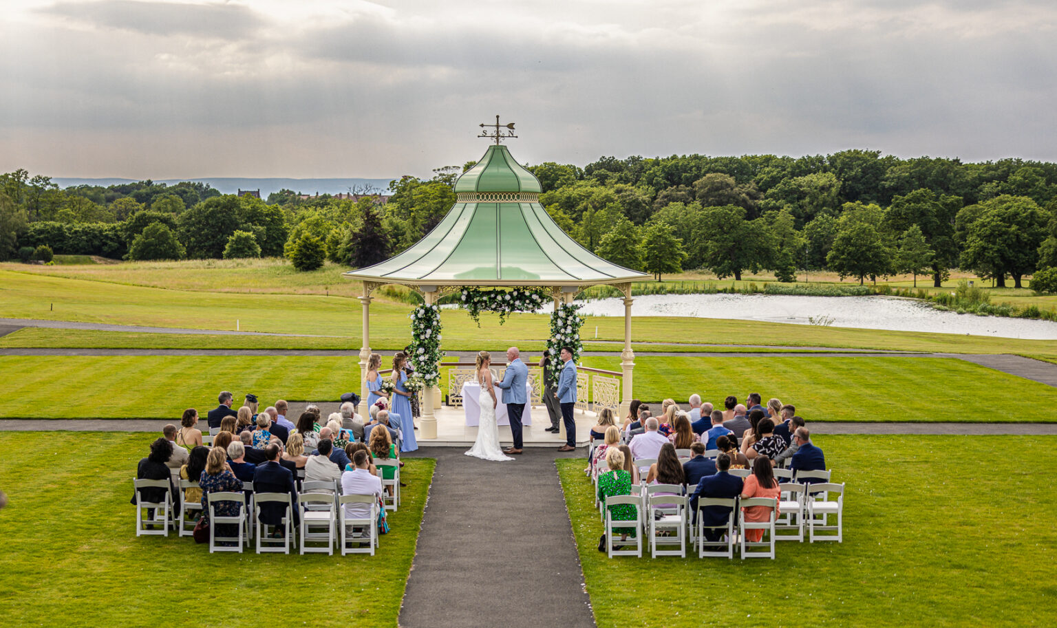 An enchanting outdoor wedding ceremony unfolds under a gazebo at Carden Park, surrounded by seated guests, with lush green grass and majestic trees setting the perfect backdrop.