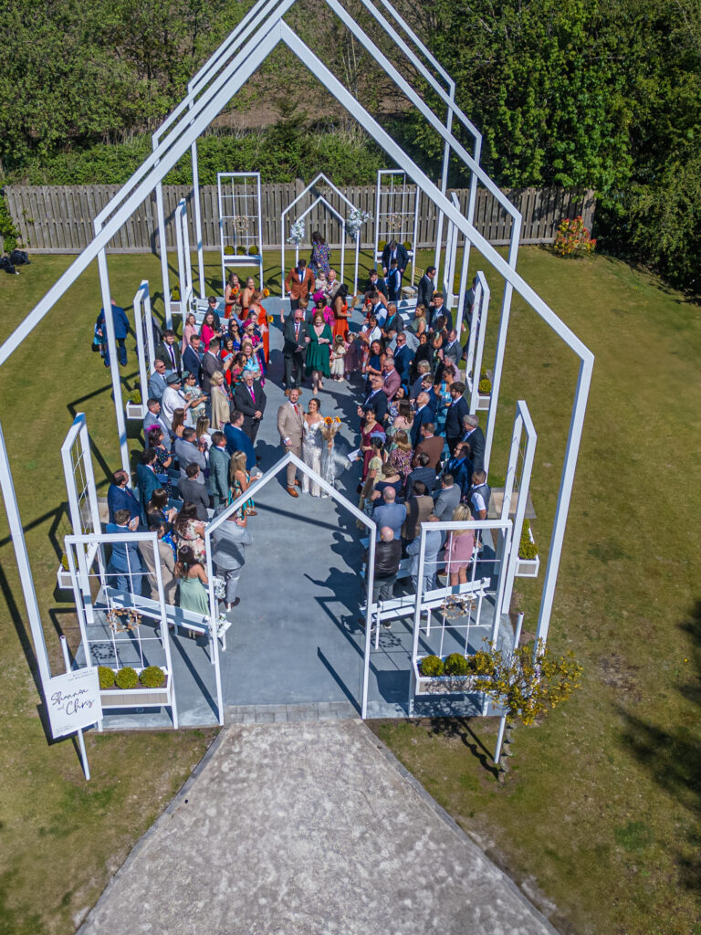 A wedding ceremony is taking place outdoors at Alcumlow Wedding Venue Cheshire, under a white open-frame structure.