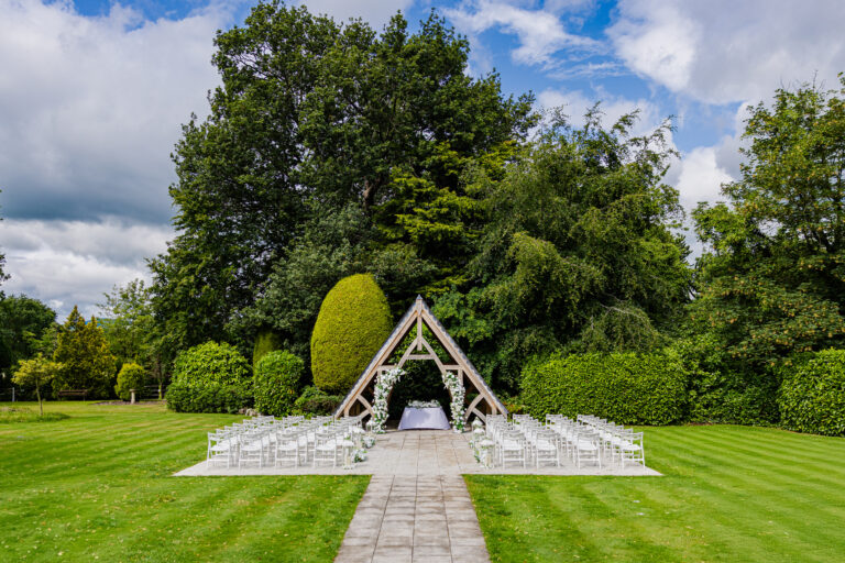 Rows of white chairs face a wooden wedding arch on a lawn, surrounded by trees and greenery under a partly cloudy sky.